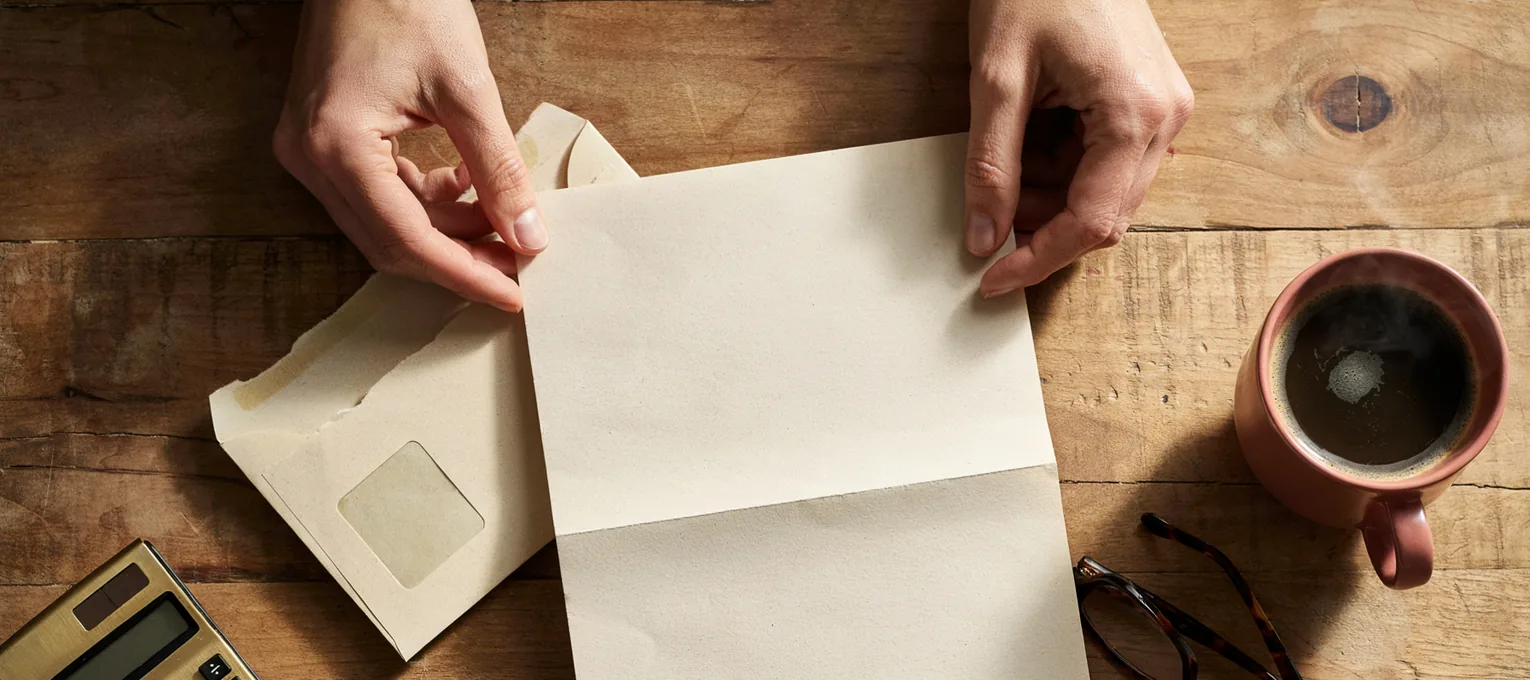 Hands opening a letter above a wooden table with a calculator and coffee — reviewing 2026 health insurance costs